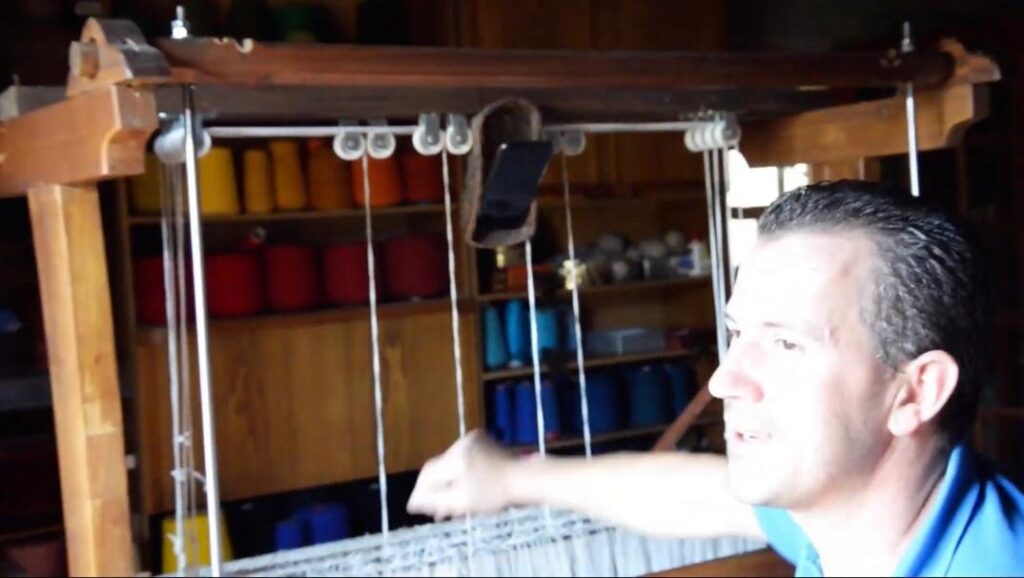 Photogramme extrait de Lá em Baixo : Fernando Rei dans son atelier à Aboim da Nóbrega, métier à tisser et smartphone suspendu.